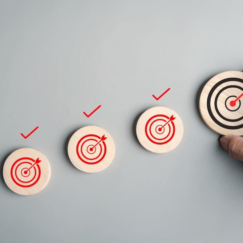 A hand places a wooden disc with a black bullseye target next to three smaller discs with red bullseyes and check marks, symbolizing progress and goal achievement.