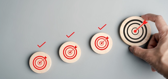 A hand places a wooden disc with a black bullseye target next to three smaller discs with red bullseyes and check marks, symbolizing progress and goal achievement.