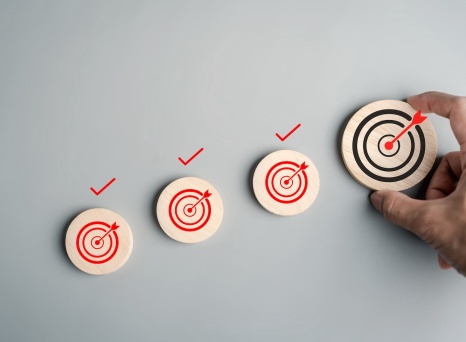 A hand places a wooden disc with a black bullseye target next to three smaller discs with red bullseyes and check marks, symbolizing progress and goal achievement.